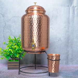 May include: A large, cylindrical, hammered copper water dispenser with a matching lid and a brass spigot. It rests on a black metal stand that includes a side holder for a matching hammered copper cup, which is also included in the set.