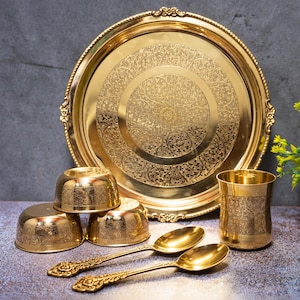 Könnte beinhalten: Ein goldfarbenes Essgeschirr-Set aus Metall, bestehend aus einem großen runden Tablett mit aufwendiger Blumengravur, drei kleinen Schalen, einem hohen Becher und zwei Löffeln. Jedes Teil weist ein passendes, detailliertes Ätzmuster auf.
