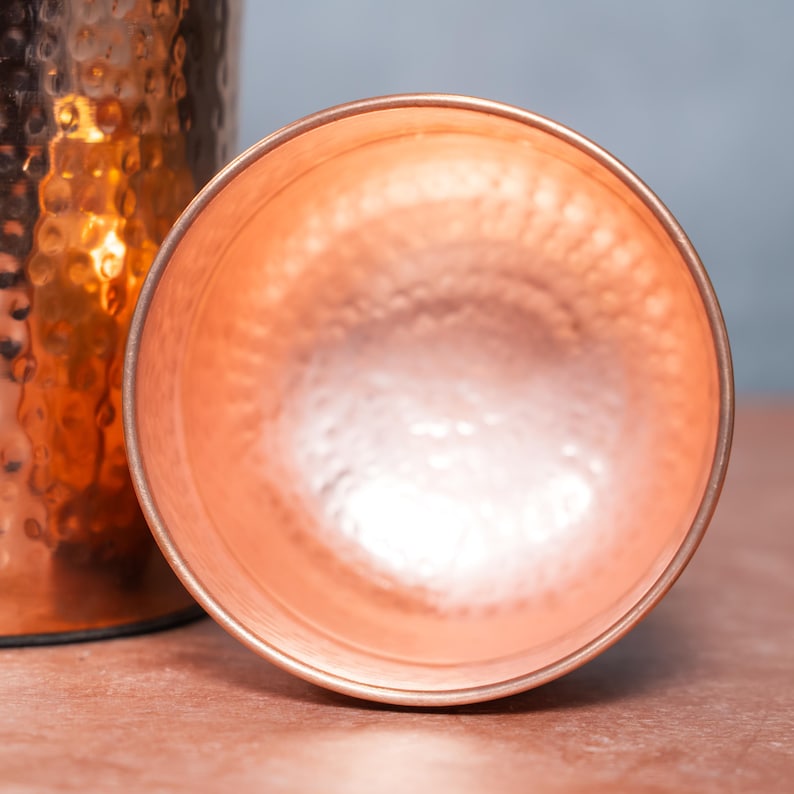 Op de afbeelding: Een bovenaanzicht van een ronde koperen beker met een duidelijke gehamerde textuur aan de binnenzijde. Het metaal heeft een warme, metaalachtige oranje tint met reflecterende accenten. Een deel van een bijpassend koperen vat is zichtbaar.