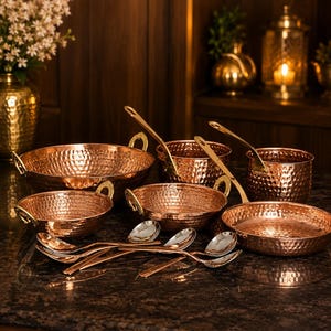 May include: A collection of hammered copper cookware including a large bowl, two smaller bowls, two deep pots, and a shallow pan, all featuring gold-toned metal handles. Several matching copper spoons are arranged in the foreground.