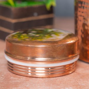 Op de afbeelding: Een ronde koperkleurige metalen deksel met een platte bovenkant voorzien van een wazig groen en geel bloemmotief. De deksel heeft een gladde, gepolijste afwerking aan de zijkanten en een witte siliconen afdichtingsring bij de schroefdraad.