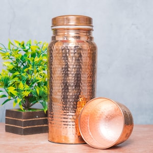 Op de afbeelding: Een cilindrische koperen waterfles met een gehamerde textuur en een bijpassend afneembaar bekerdeksel. De fles heeft een gepolijste metalen afwerking met een warme roodbruine tint, die het licht reflecteert op het oppervlak met deukjes.