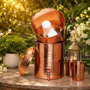 May include: A hammered copper water dispenser with a matching lid and two cylindrical copper cups. The dispenser features a metal spigot at the base. Inside the open dispenser, two white cylindrical objects are visible. The finish is metallic.