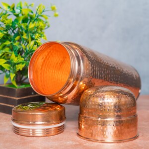 Op de afbeelding: Een cilindrische koperen waterfles met een gehamerde textuurafwerking. De fles ligt op zijn kant met de opening naar voren gericht. Twee afzonderlijke koperen doppen met een bijpassende gehamerde textuur zijn voor de fles geplaatst.