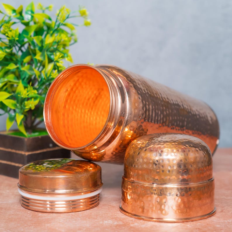 Op de afbeelding: Een cilindrische koperen waterfles met een gehamerde textuurafwerking. De fles ligt op zijn kant met de opening naar voren gericht. Twee afzonderlijke koperen doppen met een bijpassende gehamerde textuur zijn voor de fles geplaatst.