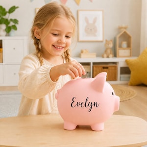 Könnte beinhalten: Ein hellrosa Sparschwein mit dem Namen Evelyn in schwarzer Schreibschrift auf der Seite. Das Sparschwein aus Keramik hat einen Schlitz auf der Oberseite zum Einwerfen von Münzen und ein kleines schwarzes Auge an der Seite des Gesichts.