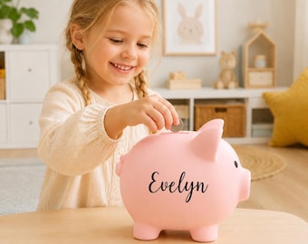 Könnte beinhalten: Ein hellrosa Sparschwein mit dem Namen Evelyn in schwarzer Schreibschrift auf der Seite. Das Sparschwein aus Keramik hat einen Schlitz auf der Oberseite zum Einwerfen von Münzen und ein kleines schwarzes Auge an der Seite des Gesichts.