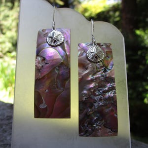 May include: A pair of rectangular dangle earrings featuring iridescent abalone shell in shades of purple, pink, and green. Each earring is topped with a small, circular silver-toned metal charm shaped like a starfish and hangs from a silver hook.