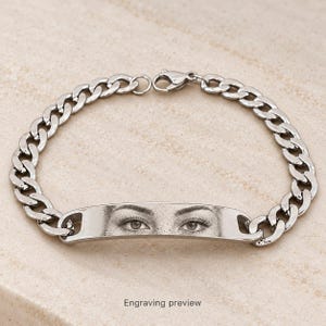 May include: A silver-toned metal curb chain bracelet featuring a central curved identification plate. The plate is engraved with a detailed black and white image of a pair of human eyes. The bracelet includes a lobster claw clasp closure.