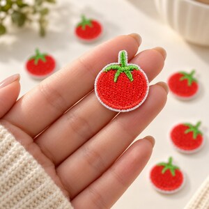 May include: A small, circular embroidered patch featuring a bright red tomato with a green stem and leaves. The patch has a white border and is shown in a collection of identical tomato-shaped patches with red and green thread.