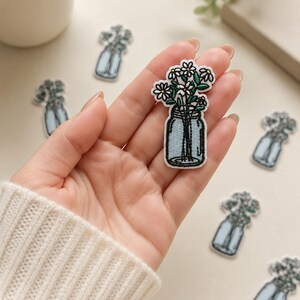May include: An embroidered patch featuring a light blue mason jar filled with white daisies and green stems. The patch has a white border and is shaped like the jar and flowers. Multiple identical patches are scattered around the main one.