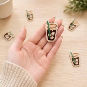May include: An embroidered patch shaped like a glass of iced coffee with a dark brown liquid, light brown ice cubes, and a bright green straw. The patch features a tan rim and is surrounded by several identical patches on a light surface.