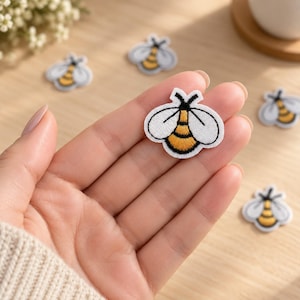May include: A small embroidered bee patch featuring white wings with black outlines and a yellow and black striped body. The patch is shaped like a bee with two antennae at the top. Several identical bee patches are scattered around the main one.