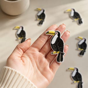 May include: A collection of embroidered toucan patches featuring a black body, a white chest, and a prominent yellow beak. Each patch is detailed with fine stitching, showcasing the bird perched on a branch, presented as a set of individual items.