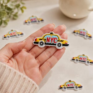 May include: An embroidered patch shaped like a yellow taxi cab with black wheels, a light blue window section, and the letters NYC stitched in red. The patch features a white border and is displayed alongside several identical taxi cab patches.
