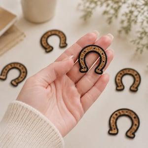 May include: Several horseshoe-shaped embroidered patches featuring a textured gold-colored center with a black stitched border and small black dots representing nail holes, arranged on a light surface with one patch held in the palm.