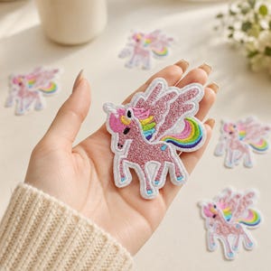 May include: An embroidered iron-on patch featuring a pink glittery unicorn with wings. The unicorn has a rainbow-colored mane and tail, with white stitched borders. Several identical unicorn patches are scattered around the central patch.