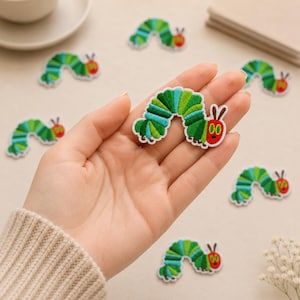 May include: An embroidered iron-on patch shaped like a green caterpillar with a red head, yellow eyes, and brown antennae. The caterpillar has a segmented body featuring various shades of green, with a white border outlining the entire shape.