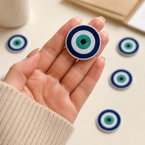 May include: A circular embroidered patch featuring a stylized eye design with a black center, a bright green iris, a white ring, and a dark blue outer border. The patch is held in a palm, with several identical patches scattered nearby on a surface.