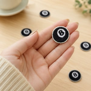 May include: A circular embroidered patch featuring a black billiard eight ball design with a white border and a white number eight in the center. The patch is small and round, with detailed stitching that creates a textured surface on the fabric.