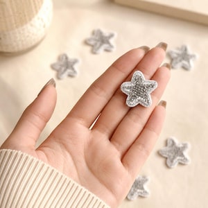 May include: A small, five-pointed star patch featuring metallic silver embroidery with a white stitched border. The star is held in the palm, while several identical silver star patches are scattered around on a light-colored surface.