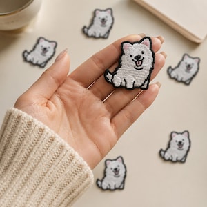 May include: A white embroidered dog patch with black outlines, featuring a sitting puppy with a happy expression, pink inner ears, and a black nose. The patch is shown in a hand, with several identical dog patches scattered around the area.
