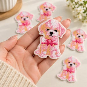 May include: An embroidered patch featuring a puppy with light pink and golden yellow fur, wearing a bright pink bow around its neck. The puppy has black eyes and a black nose, with a white border outlining the entire shape of the dog figure.