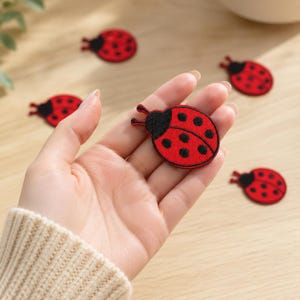 May include: Multiple embroidered ladybug patches featuring a bright red body with black spots and a black head with two antennae. Each patch is circular in shape with a distinct black border, showcasing a detailed insect design for textile application.