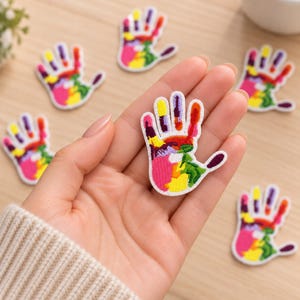 May include: A set of embroidered patches shaped like handprints, each featuring a vibrant, multi-colored design with segments of pink, yellow, green, orange, purple, and red. The patches have a white border and a textured, stitched surface.
