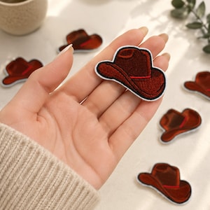 May include: A collection of embroidered iron-on patches shaped like cowboy hats. Each patch features a deep brown color with a thin red accent line along the brim and a white border. The patches are small, detailed, and have a textured finish.