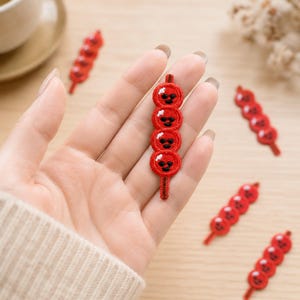May include: A red embroidered patch shaped like a skewer of four round dango dumplings, each featuring a small black stitched face and a white highlight. The patch is vertically oriented with a thin red stem at the bottom and top, showcasing detailed thread work.