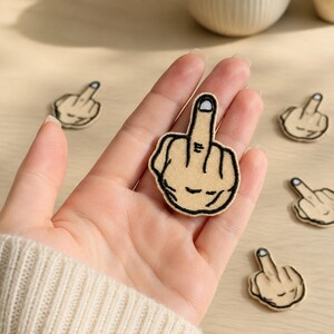 May include: A beige embroidered patch featuring a hand gesture with the middle finger extended. The design is outlined in thick black thread, with a white fingernail detail at the tip of the finger. The patch has a tan fabric base.