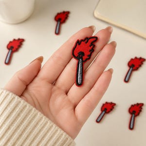 May include: A small embroidered patch shaped like a lit matchstick with a bright red flame and a grey stem outlined in black thread. The patch features a textured fabric surface and is shown against a neutral background with several identical pieces.