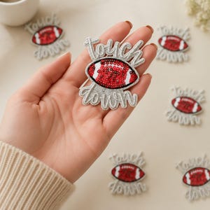 May include: A sequined patch featuring a red and white American football design with the word touchdown embroidered in silver script lettering. The patch is held in a hand, with several identical patches scattered in the background.
