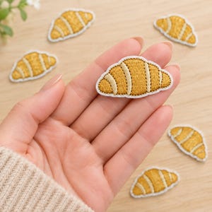May include: A collection of small, embroidered croissant patches featuring golden-yellow thread with light beige accent lines. Each patch has a distinct crescent shape with a white border, showcasing detailed stitching that mimics the layers of a pastry.