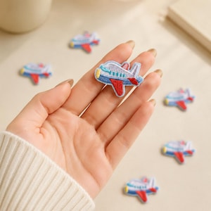 May include: A small embroidered airplane patch featuring a light blue body, a red wing, and a yellow nose. The patch has a white outline and is designed with stitched details representing windows. Several identical airplane patches are scattered nearby.