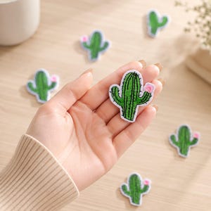 May include: A green cactus-shaped embroidered patch with a small pink flower detail on the side. The patch features white stitched borders and dark green vertical lines representing the cactus texture, held in a palm against a light background.