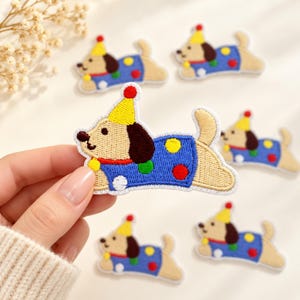 May include: An embroidered patch featuring a tan dog with brown ears wearing a yellow party hat and a blue shirt decorated with colorful red, green, yellow, and white polka dots. The dog is depicted in a playful, horizontal pose.