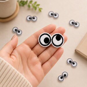 May include: A small, embroidered patch featuring two cartoon eyes with white sclera, black pupils, and thick black outlines. The patch is oval-shaped, showing two circular eyes joined together, with one pupil looking left and the other looking right.
