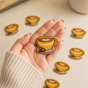 May include: An embroidered patch featuring a yellow coffee cup with a white heart-shaped latte art design, a brown saucer, and two coffee beans. The patch is circular in shape with a dark brown border, showcasing detailed stitching and vibrant colors.