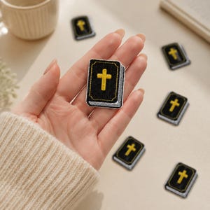 May include: A small rectangular embroidered patch shaped like a book with a black cover and a yellow cross centered on the front. The patch features white and black stitching along the edges to represent pages, with several identical patches nearby.