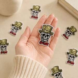 May include: An embroidered patch featuring a skull wearing a top hat with a green dollar bill pattern and a red neckerchief. The skull is white with black details, and the patch has a white border. Multiple identical patches are scattered around.