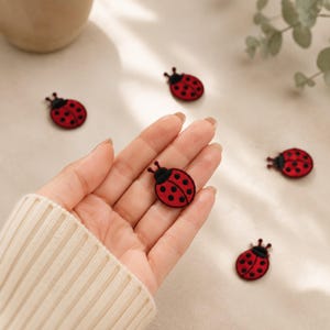 May include: Small ladybug-shaped patches featuring a vibrant red body with black spots and a black head. These circular embroidered embellishments are designed with a flat surface and detailed stitching, showcasing a classic insect motif in a compact size.