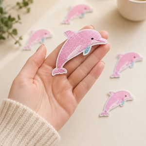 May include: A pink dolphin embroidered patch with a white border and a small teal accent on the fin. The patch features a stitched texture and a black eye detail. Several identical dolphin patches are scattered around the central piece.