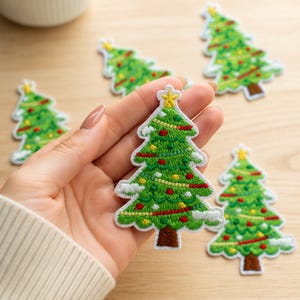 May include: A set of embroidered Christmas tree patches featuring a green tree with a brown trunk, a yellow star topper, and colorful red and yellow ornaments. The patches have white accents representing snow on the branches and a white border.