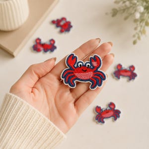 May include: A small, embroidered iron-on patch shaped like a red crab with dark blue outlines, white eyes with black pupils, and a light pink belly. The crab has two raised claws and six legs, featuring a stitched texture throughout the design.