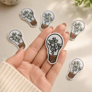 May include: Multiple embroidered patches shaped like light bulbs, each featuring a white background with a black outline and a brown base. Inside each bulb is a green plant with small leaves. One patch is held in the palm of a hand.