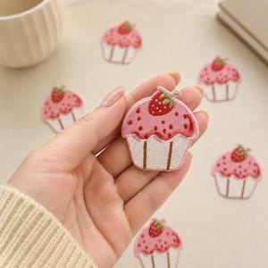 May include: An embroidered patch shaped like a cupcake with pink frosting, red strawberry accents, and a white base. The cupcake features a small red strawberry on top, dark pink dots, and brown vertical lines on the white bottom section of the patch.
