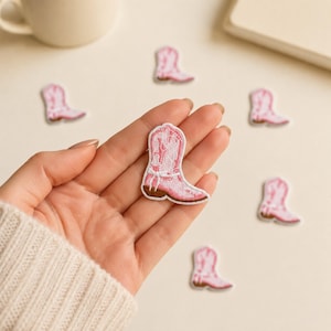 May include: A collection of small, embroidered iron-on patches shaped like cowboy boots. Each patch features a light pink color with white stitching details, a white decorative harness strap, and a brown sole. The patches are shown in a group.