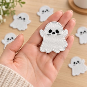 May include: A white embroidered ghost patch with black stitched eyes and mouth, featuring small grey accents under the eyes. The patch has a rounded top and a scalloped bottom edge, with a white border surrounding the entire ghost shape.